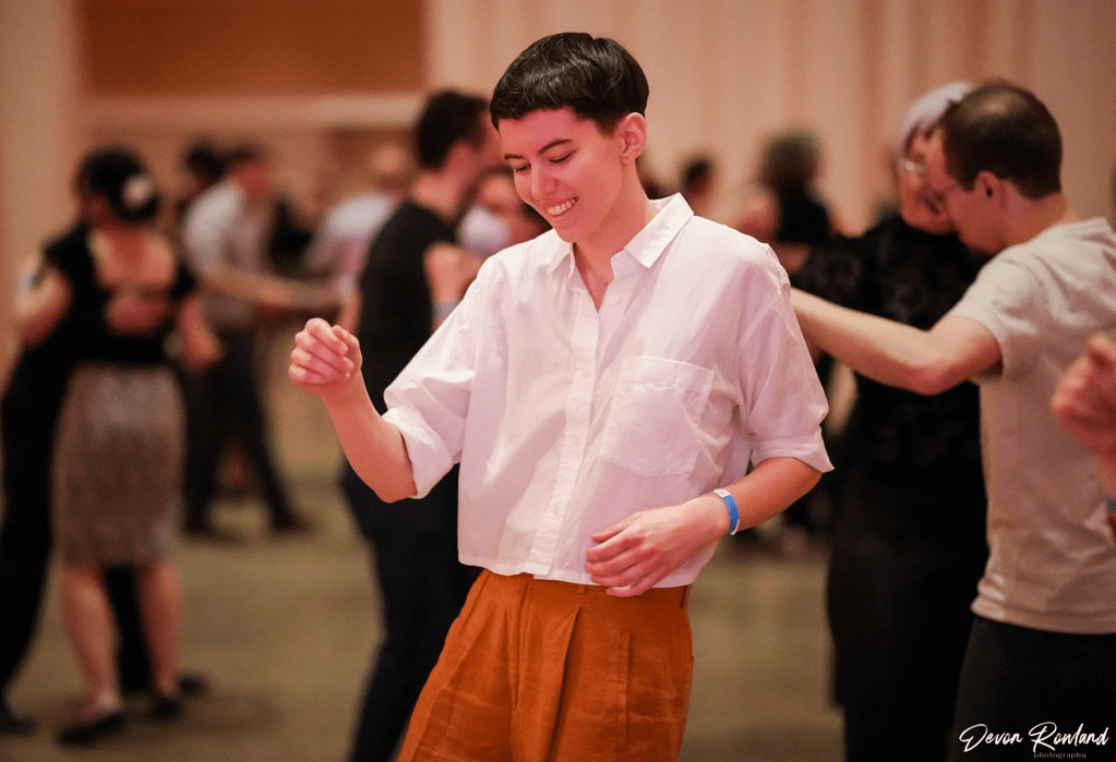 Mary, a white person with short brown hair wearing a white shirt and orange pants, is dancing by themselves and smiling.